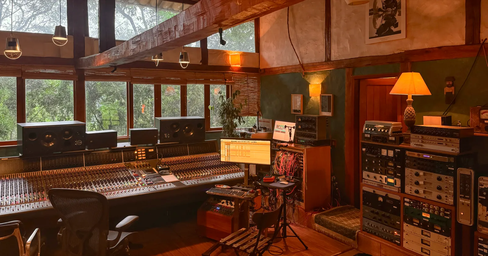 A control room with a large mixing console, studio monitors in front of tall windows onto green trees, and racks of outboard gear