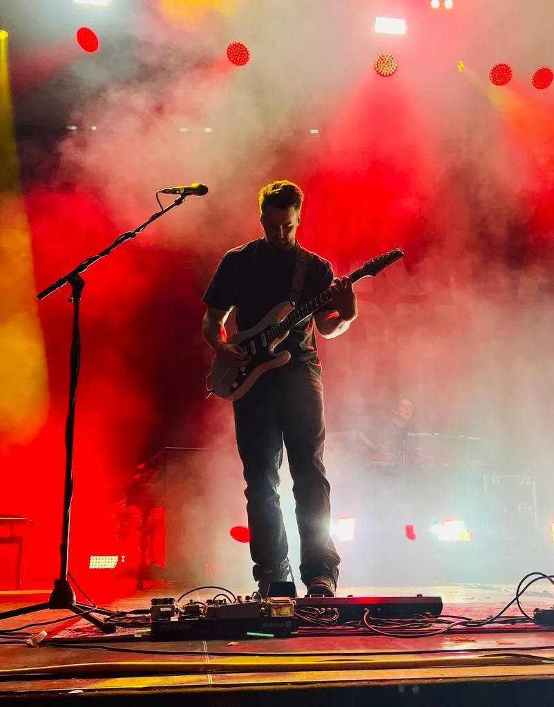 A guitarist on a smoky, red-lit stage, a microphone stand and pedalboard in front of him